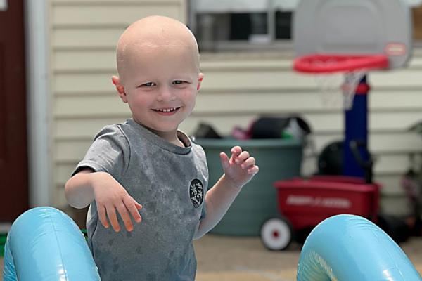A young boy smiles while standing behind a backyard water slide. In the background, a box of toys and a children’s basketball hoop sit on the porch in front of a home. The door and windows are visible.