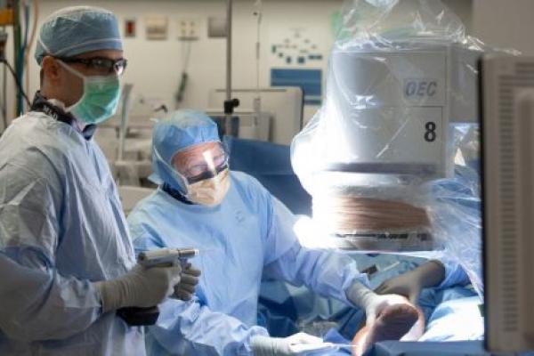 Medical professionals in full surgical attire perform a procedure in an operating room. One holds a surgical drill while another works on a patient's leg.
